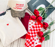 Adult tee in white at the top of a stack with mini symbol heart design on front, shown on flat lay with other valentines props surrounding – Little Mama Shirt Sho