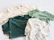 Green and ivory adult CC tees with lucky charm marshmallows embroidered on chest and sage green child tee and natural child sweater with printed lucky charm marshmallows, seen on flat lay - Little Mama Shirt Shop