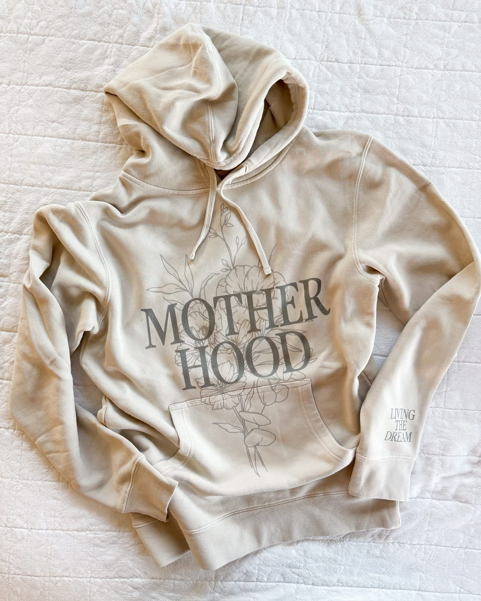 Adult pigment hoodie in cream with 'motherhood' printed on front with floral design behind and 'living the dream' printed on the sleeve, seen on flat lay - Little Mama Shirt Shop