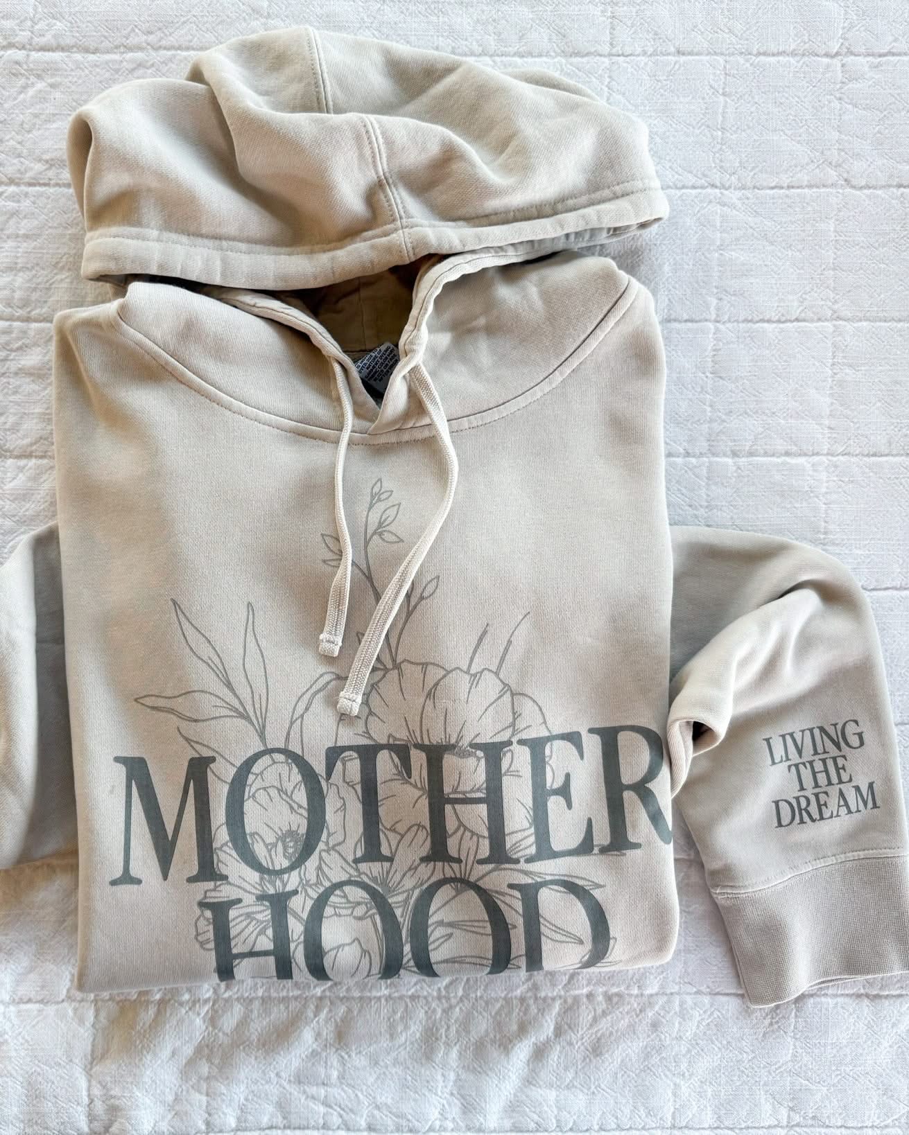 Adult pigment hoodie in cream with 'motherhood' printed on front with floral design behind and 'living the dream' printed on the sleeve, seen on flat lay - Little Mama Shirt Shop