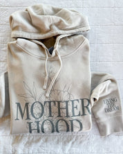 Adult pigment hoodie in cream with 'motherhood' printed on front with floral design behind and 'living the dream' printed on the sleeve, seen on flat lay - Little Mama Shirt Shop