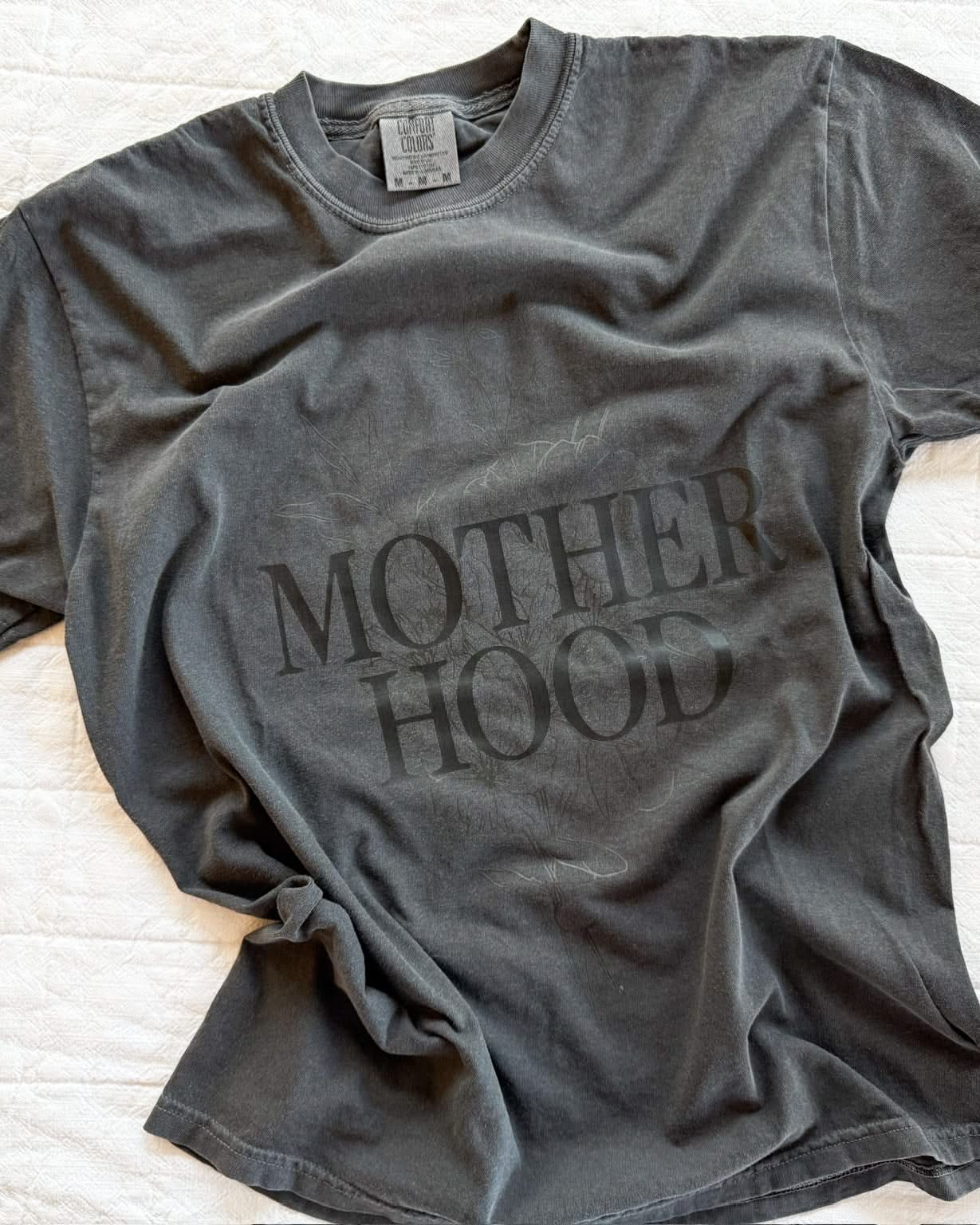 Adult tee in pepper with 'motherhood' printed on the front with floral design behind, seen on flat lay - Little Mama Shirt Shop