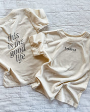 Child tee in cream with 'boyhood' printed on the front and 'this is the good life' printed on the back, seen on flat lay - Little Mama Shirt Shop