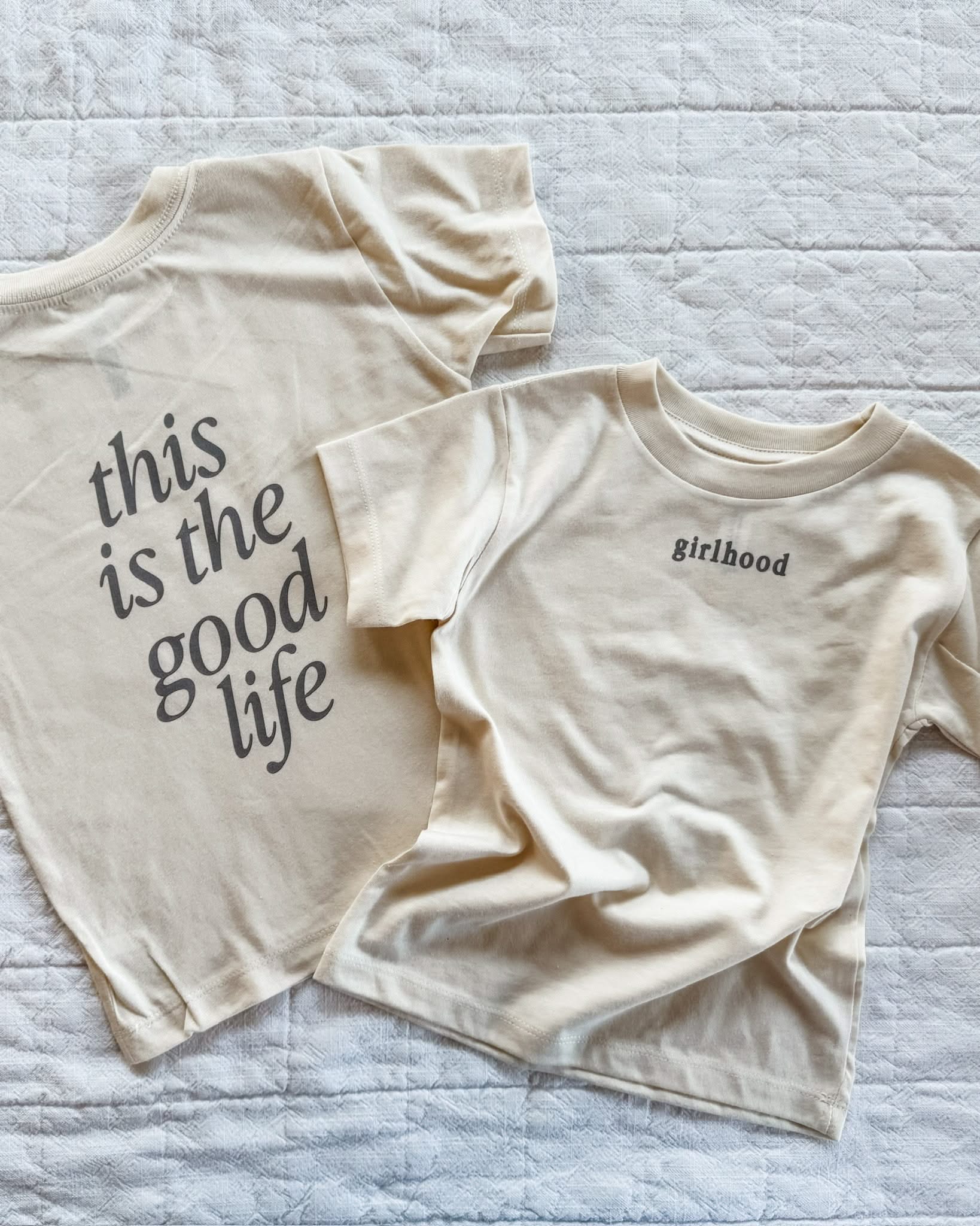 Child tee in cream with 'girlhood' printed on the front and 'this is the good life' printed on the back, seen on flat lay - Little Mama Shirt Shop