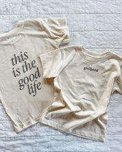 Child tee in cream with 'girlhood' printed on the front and 'this is the good life' printed on the back, seen on flat lay - Little Mama Shirt Shop