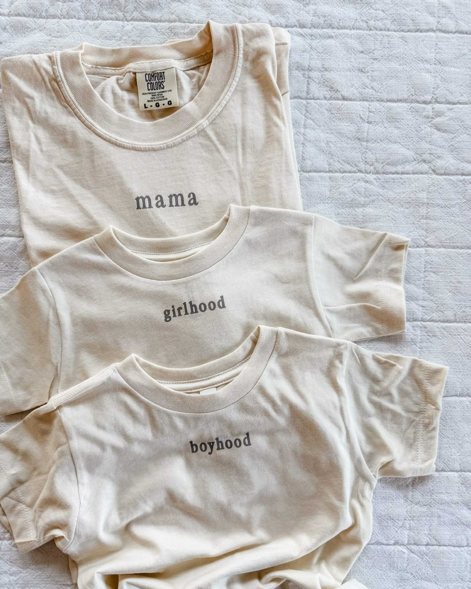 Adult tee in ivory and child tee in cream with 'mama', 'girlhood', and 'boyhood' printed on the front, seen on flat lay - Little Mama Shirt Shop