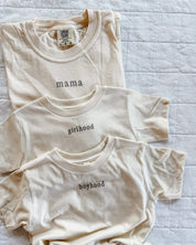 Adult tee in ivory and child tee in cream with 'mama', 'girlhood', and 'boyhood' printed on the front, seen on flat lay - Little Mama Shirt Shop