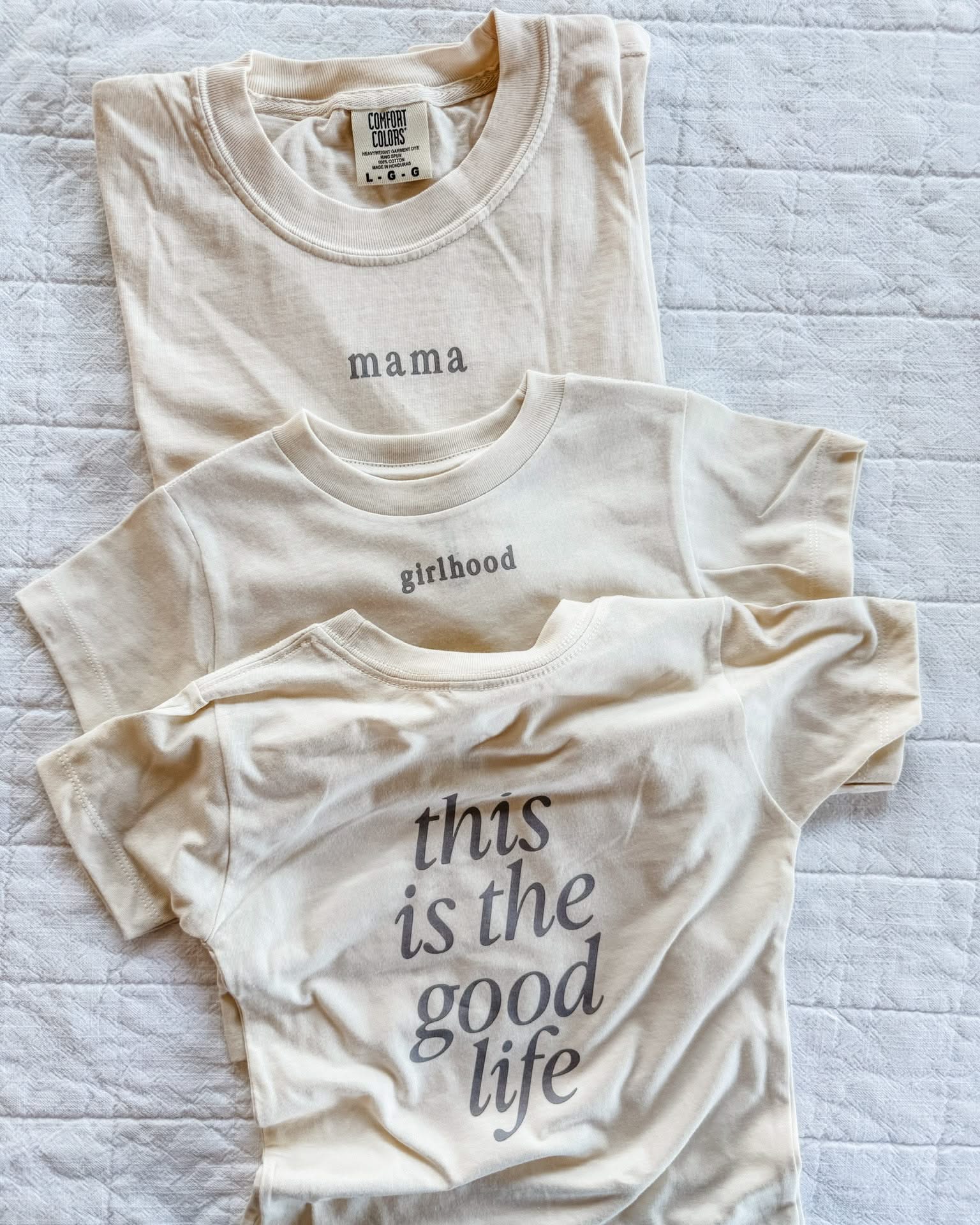 Adult tee in ivory and child tee in cream with 'mama' and 'girlhood' printed on the front and 'this is the good life' printed on back, seen on flat lay - Little Mama Shirt Shop