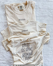 Adult tee in ivory and child tee in cream with 'mama' and 'girlhood' printed on the front and 'this is the good life' printed on back, seen on flat lay - Little Mama Shirt Shop