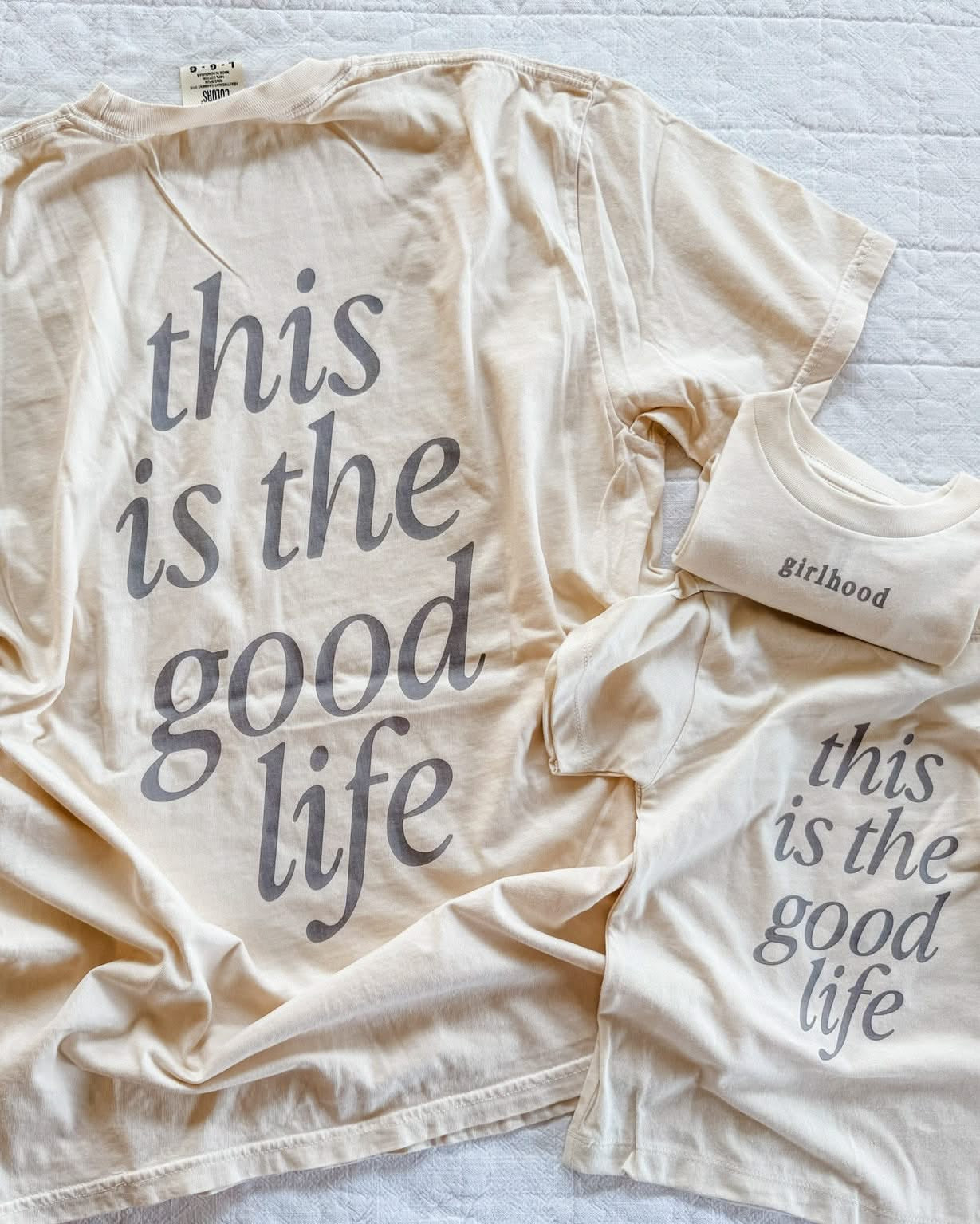 Adult tee in ivory and child tee in cream with 'girlhood' printed on the front and 'this is the good life' printed on the back, seen on flat lay - Little Mama Shirt Shop