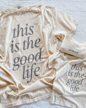 Adult tee in ivory and child tee in cream with 'girlhood' printed on the front and 'this is the good life' printed on the back, seen on flat lay - Little Mama Shirt Shop