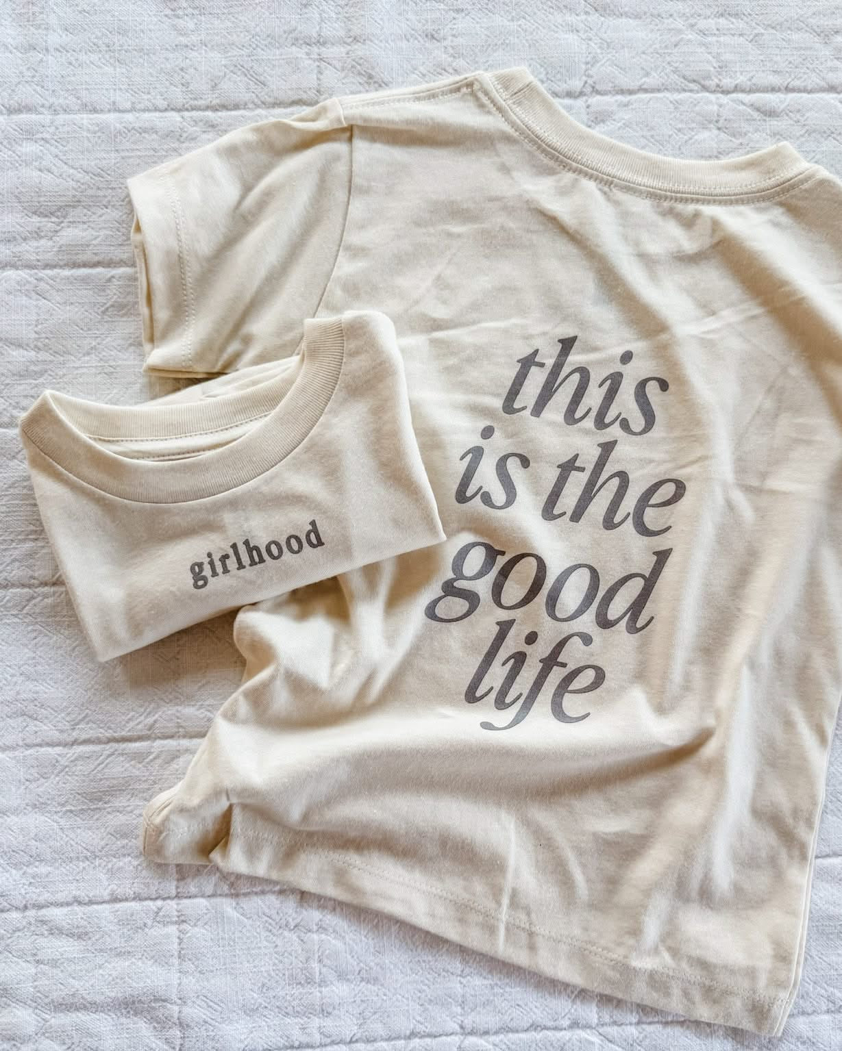 Child tee in cream with 'girlhood' printed on the front and 'this is the good life' printed on the back, seen on flat lay - Little Mama Shirt Shop