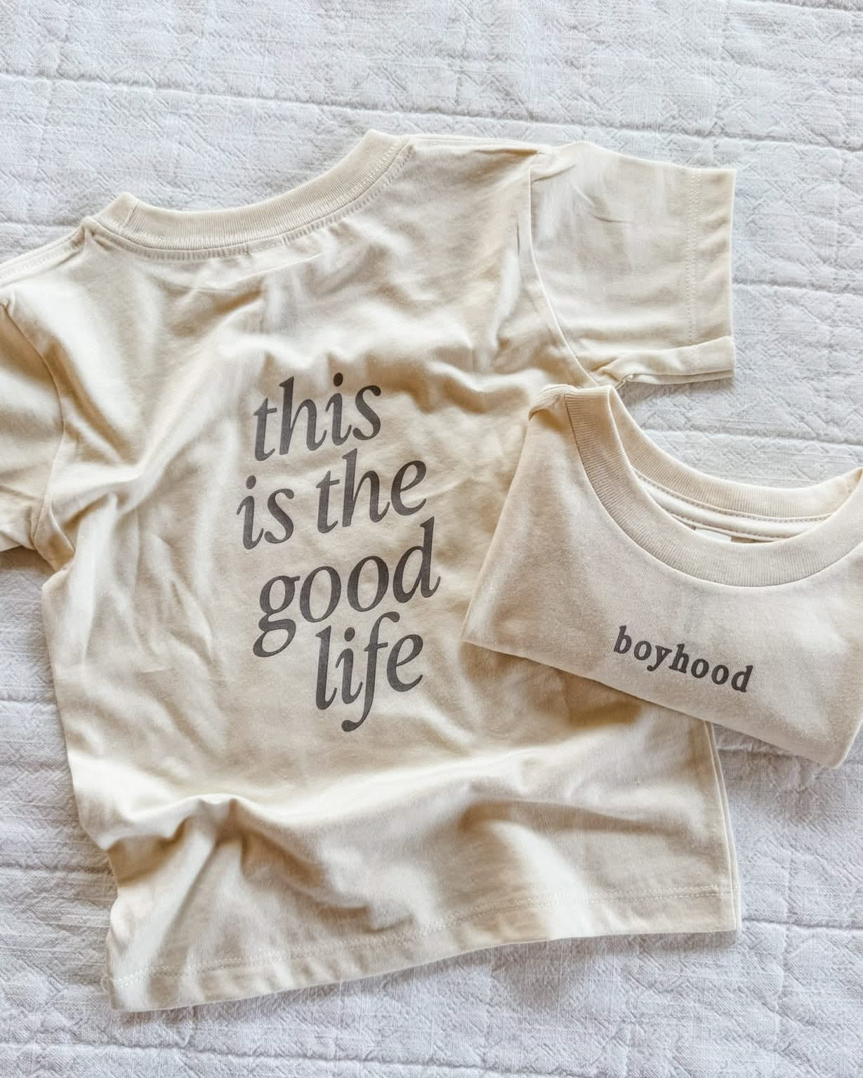 Child tee in cream with 'boyhood' printed on the front and 'this is the good life' printed on the back, seen on flat lay - Little Mama Shirt Shop