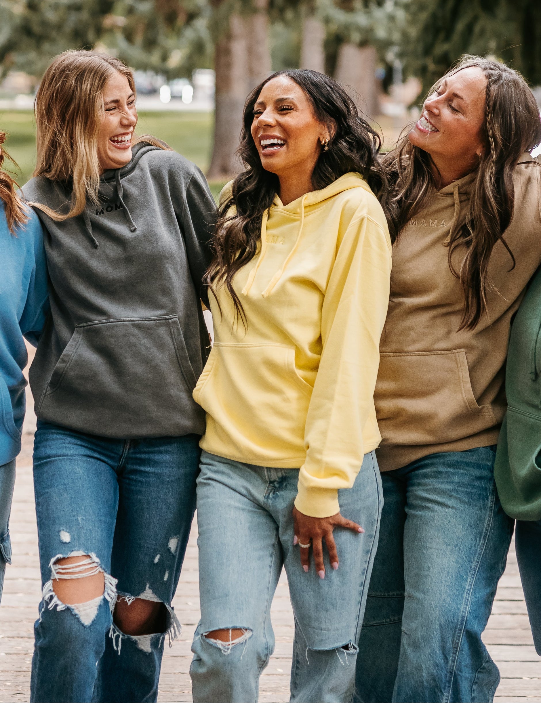 Five women in colorful hoodies and jeans standing together outdoors with trees in the background.