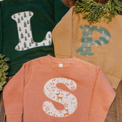 Child sweaters in forest, coyote brown, and mauvelous with Christmas patterned initials on front, shown on flat lay – Little Mama Shirt Shop