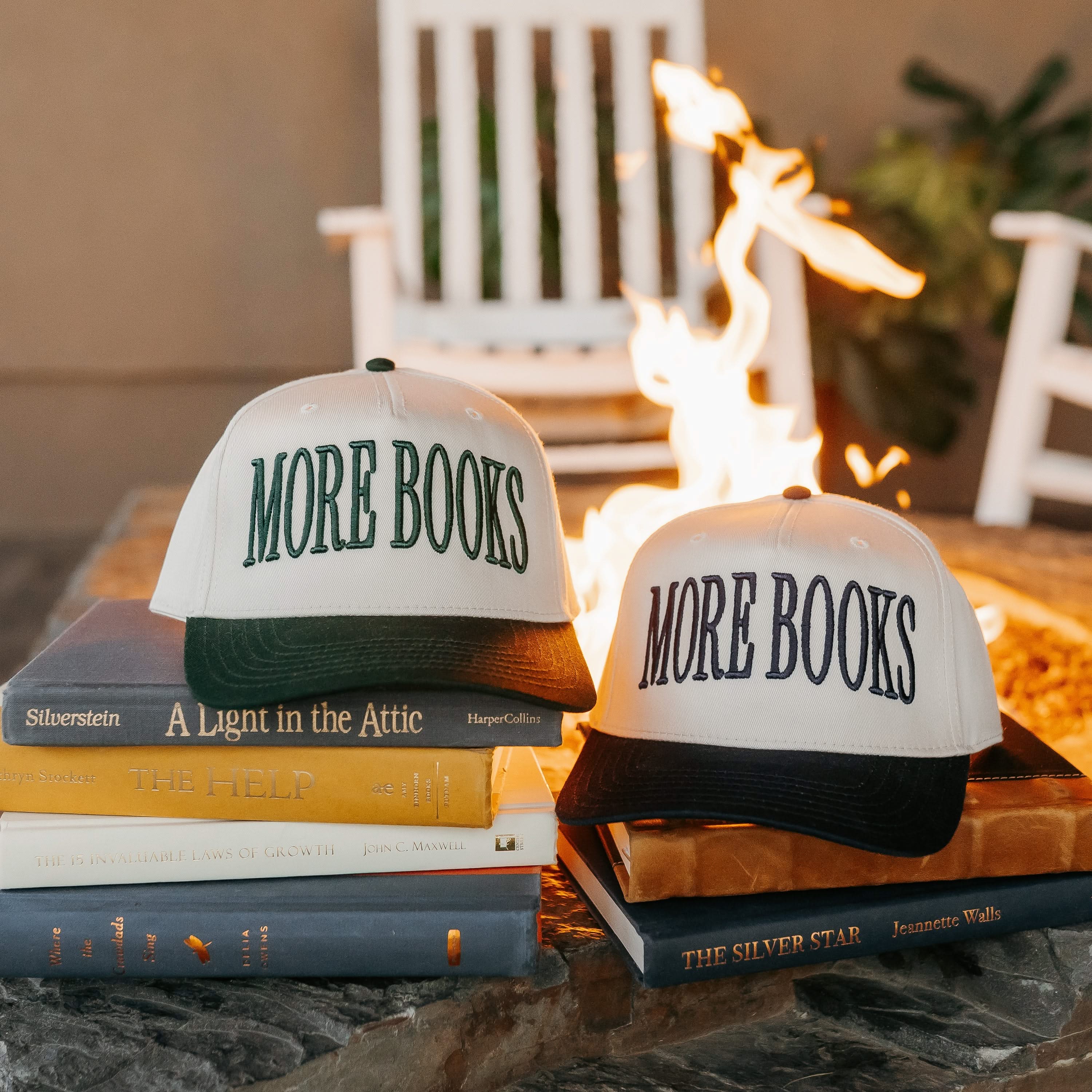 Tan and green and tan and black trucker hats, featuring a front embroidered design that reads “More Books" sitting on books by a fire. - Little Mama Shirt Shop