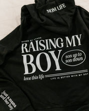 Hoodie in black with white 'Raising My Boy' graphic on flat lay - Little Mama Shirt Shop