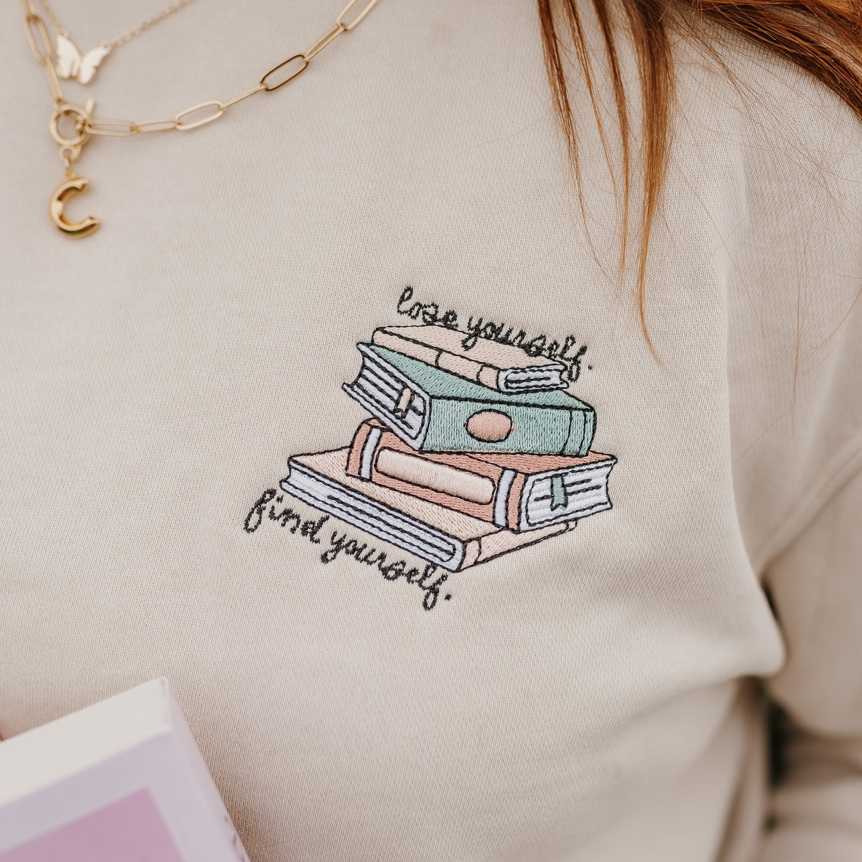 Close up unisex “Lose Yourself, Find Yourself” sweatshirt in pigment sand with embroidered book stack on model – Little Mama Shirt Shop