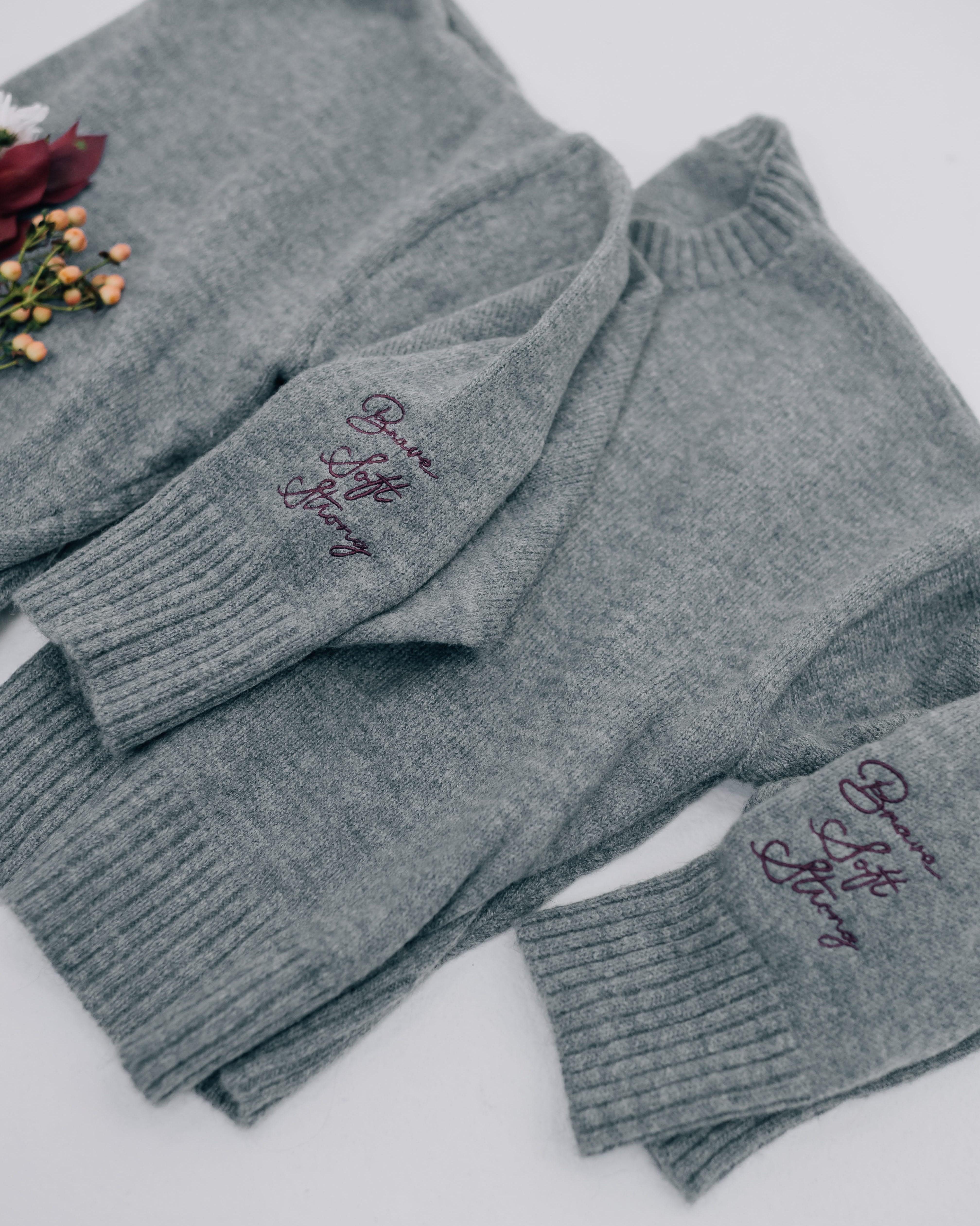 Gray sweaters with "Brave, Soft, Strong" embroidered on the sleeve, seen on flat lay - Little Mama Shirt Shop