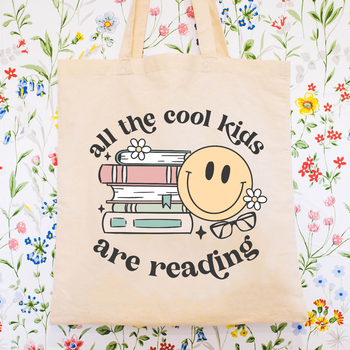 Beige canvas tote bag featuring a 'all the cool kids are reading' design on a flat lay - Little Mama Shirt Shop