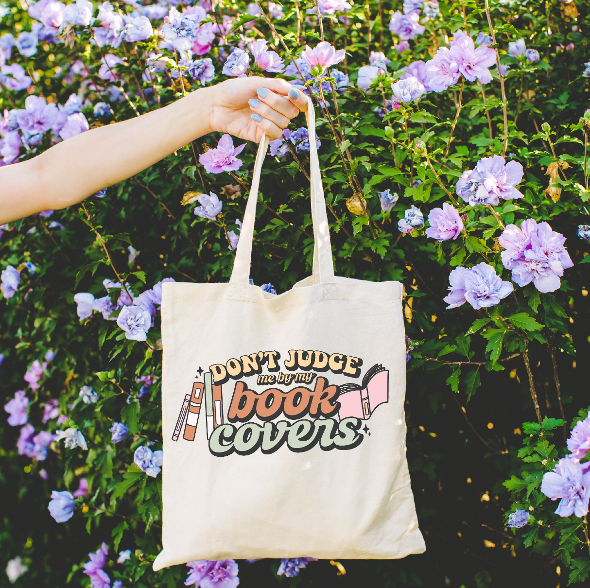 Beige canvas tote bag featuring a 'Don't Judge me by my Book Covers' design on a flat lay - Little Mama Shirt Shop