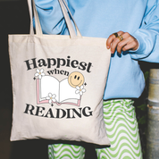 Beige canvas tote bag featuring a 'Happiest when Reading' design on a model - Little Mama Shirt Shop