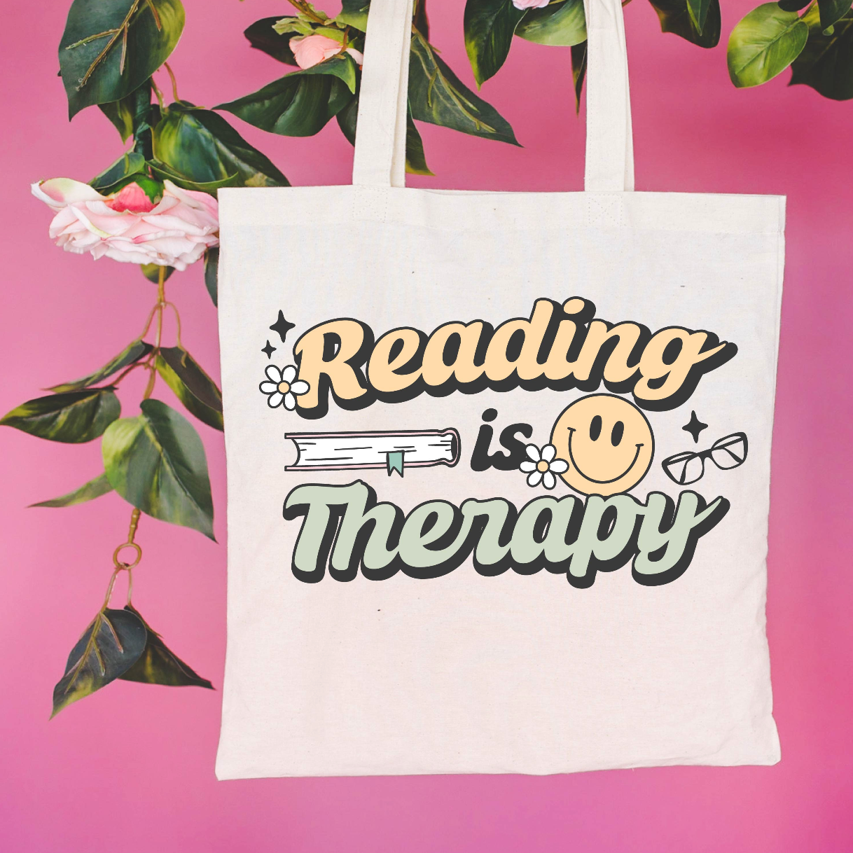 Beige canvas tote bag featuring a 'Reading is Therapy' design on a flat lay - Little Mama Shirt Shop
