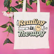 Beige canvas tote bag featuring a 'Reading is Therapy' design on a flat lay - Little Mama Shirt Shop