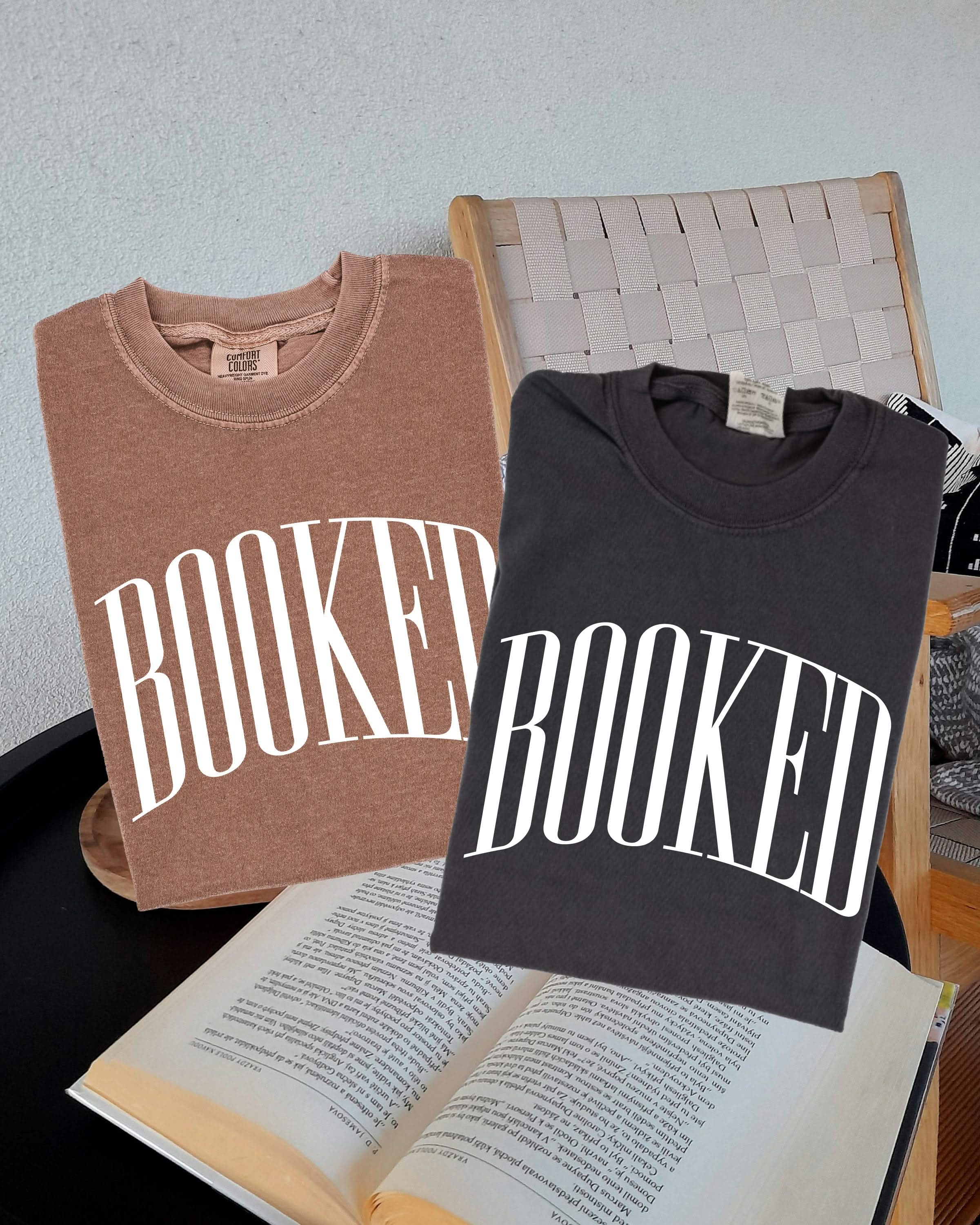 2 comfort colors tees in espresso and pepper with 'BOOKED' graphic on chest, shown on flat lay – Little Mama Shirt Shop
