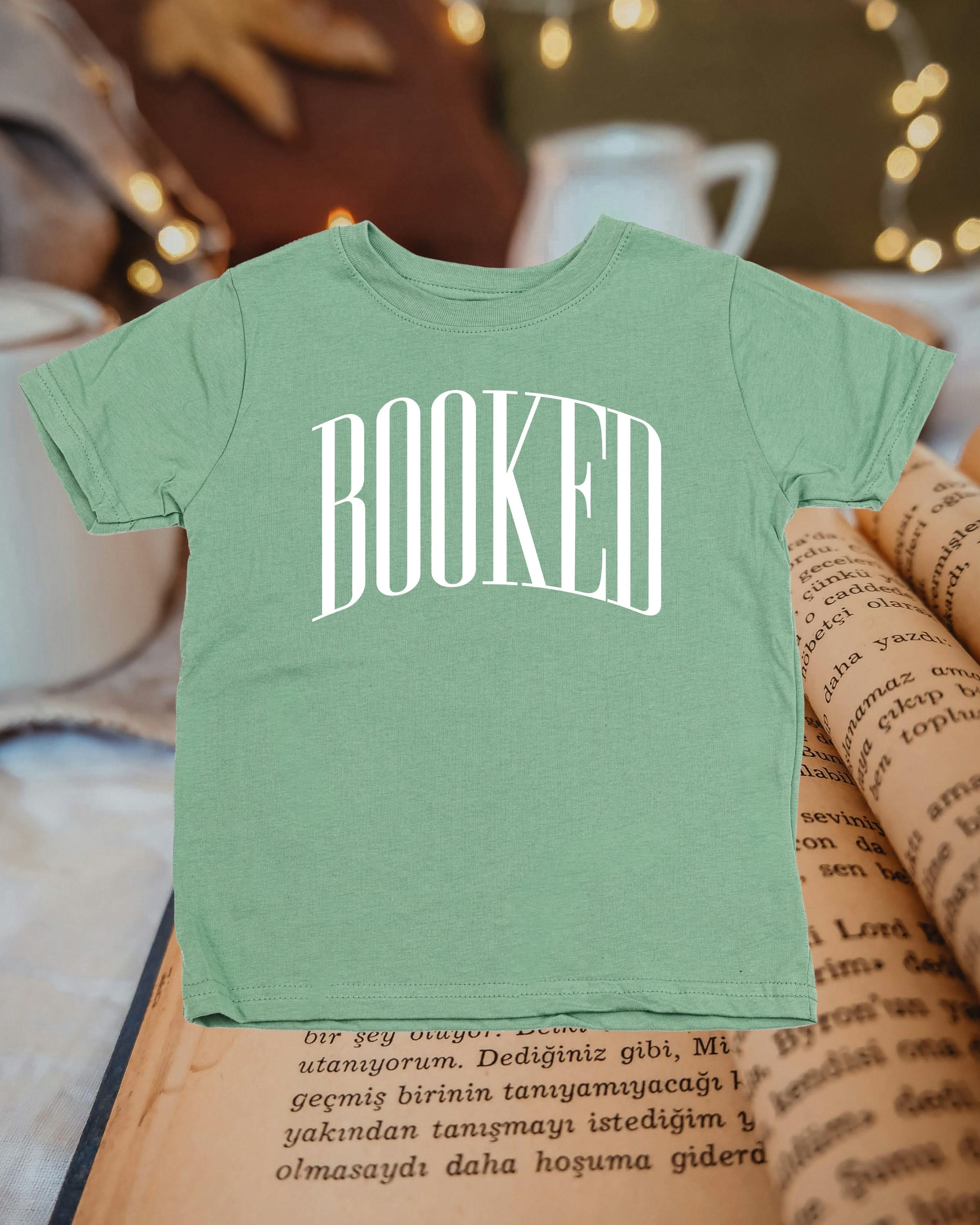 Child tee in sage with 'BOOKED' graphic on chest, shown on flat lay – Little Mama Shirt Shop