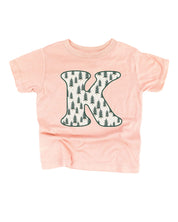 Child tee in blush with Christmas pattern trees with initial K on front, shown on flat lay – Little Mama Shirt Shop