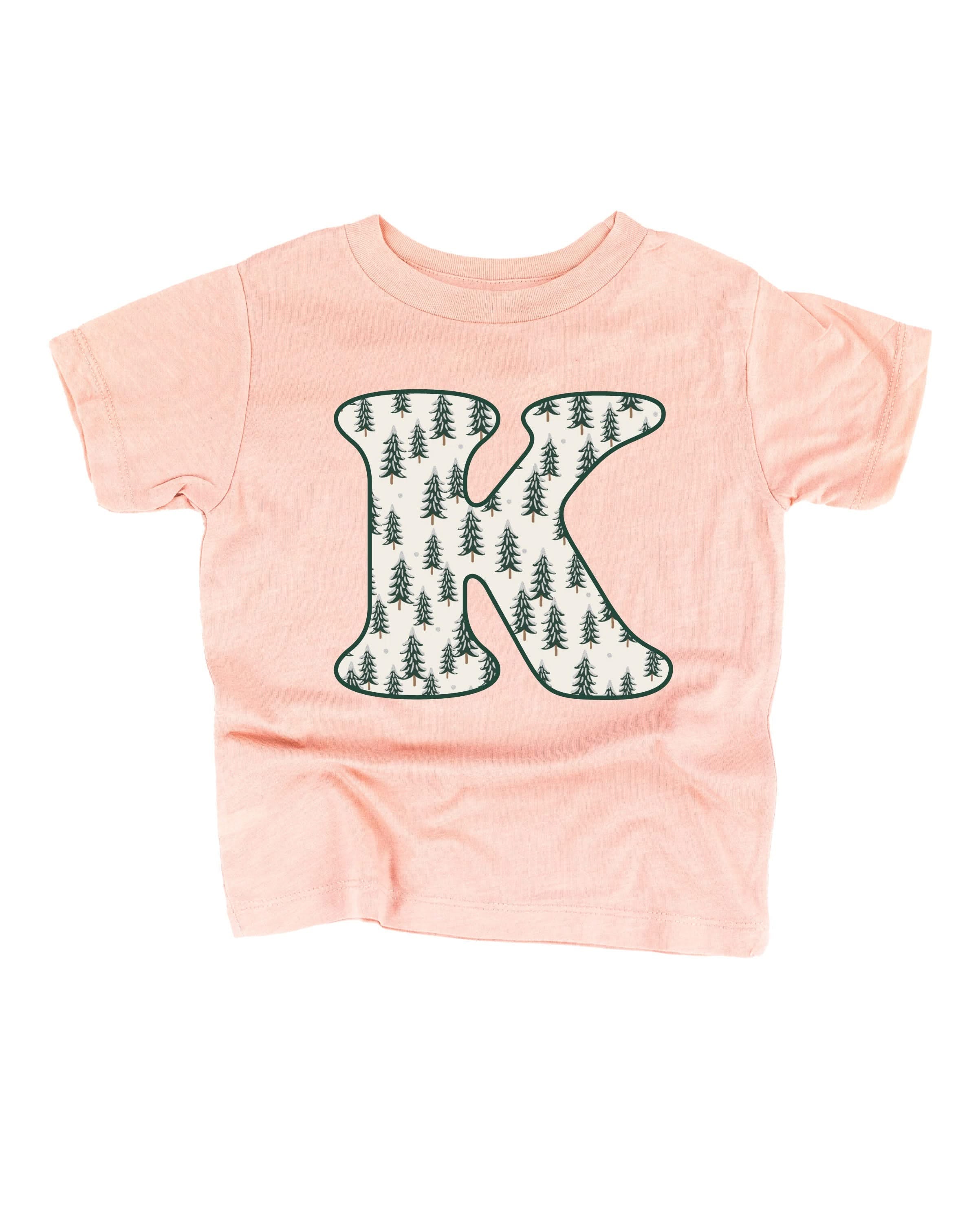 Child tee in blush with Christmas pattern trees with initial K on front, shown on flat lay – Little Mama Shirt Shop