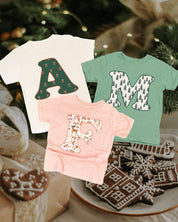 Child tees in natural, sage, and blush with Christmas patterned initials on front, shown on flat lay – Little Mama Shirt Shop