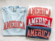 Folded AMERICA shirts with gingham lettering in red, white, and blue, showcasing a clean and simple display