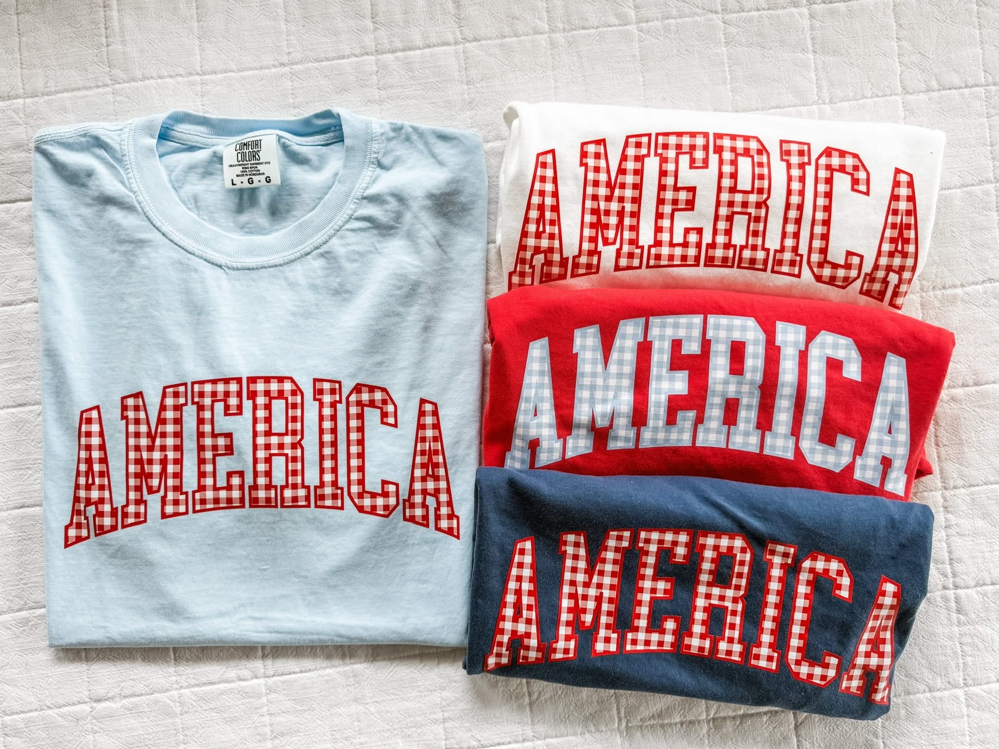 Folded AMERICA shirts with gingham lettering in red, white, and blue, showcasing a clean and simple display