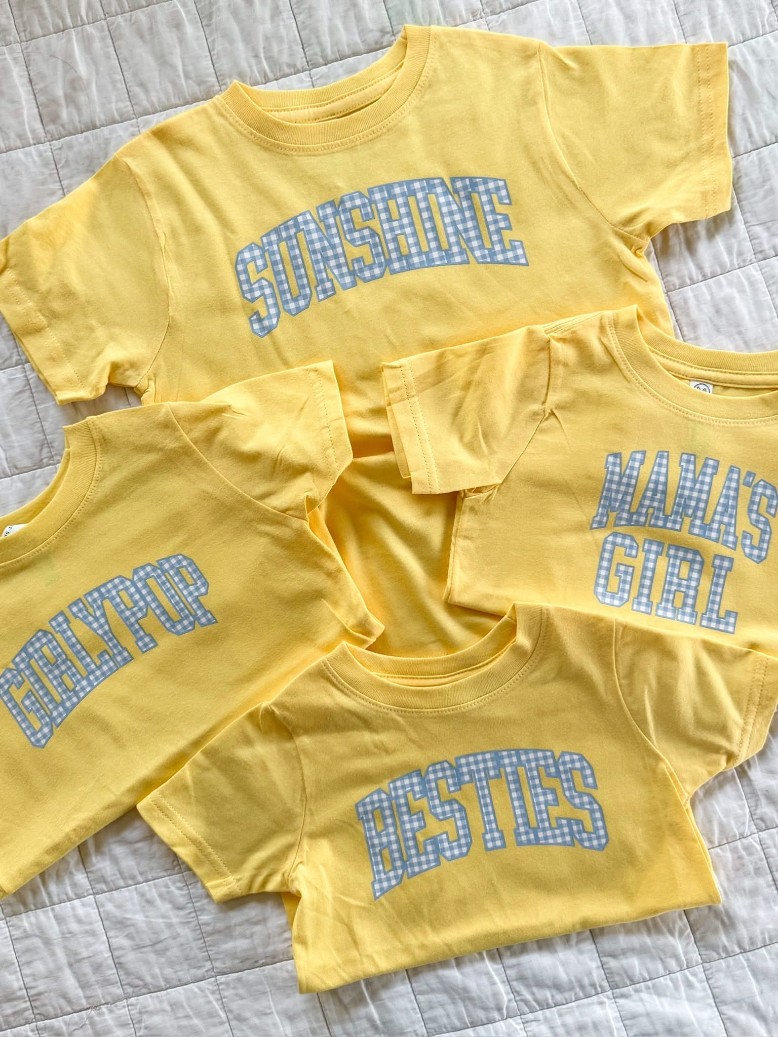 Stack of butter yellow girls tees with blue gingham lettering featuring fun phrases for kids