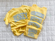 Butter yellow girls shirts with blue gingham phrases including honey, besties, girlfriend, and mama’s girl laid out together