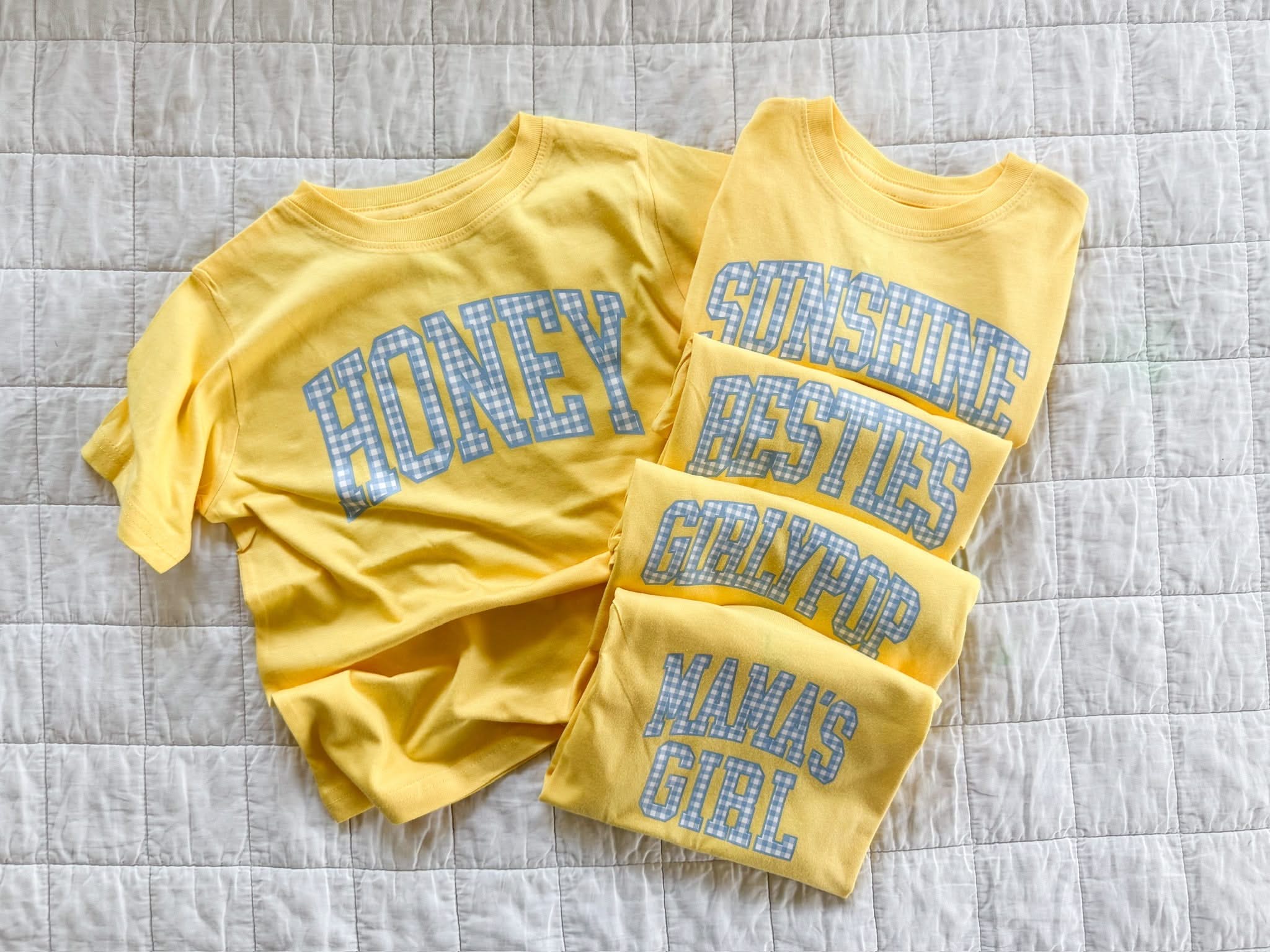 Butter yellow girls shirts with blue gingham phrases including honey, besties, girlfriend, and mama’s girl laid out together