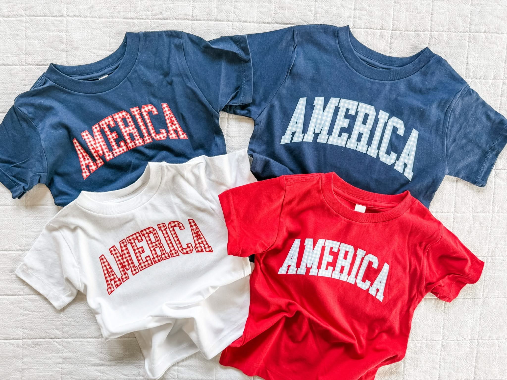 Kids AMERICA shirts in red, white, and blue with gingham lettering, laid out together for a classic summer look