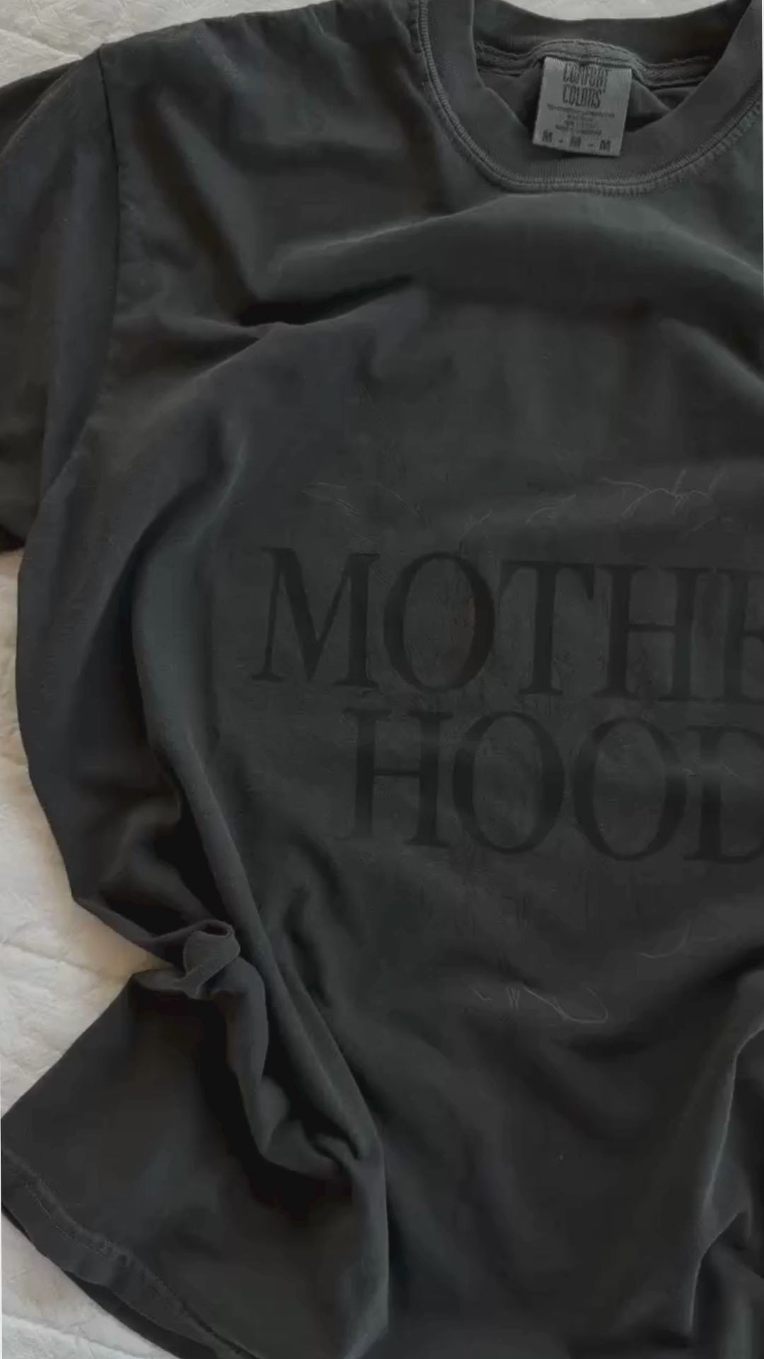 Video of adult tee in pepper with 'motherhood' printed on the front with floral design behind, seen on flat lay - Little Mama Shirt Shop