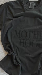 Video of adult tee in pepper with 'motherhood' printed on the front with floral design behind, seen on flat lay - Little Mama Shirt Shop