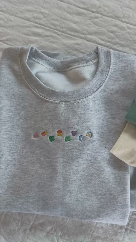Video of ash adult basic fleece and green and ivory CC tees with lucky charm marshmallows embroidered on chest, seen on flat lay - Little Mama Shirt Shop
