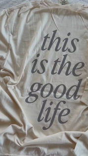 Video of adult tee in ivory and child tee in cream with 'boyhood' printed on the front and 'this is the good life' printed on the back, seen on flat lay - Little Mama Shirt Shop