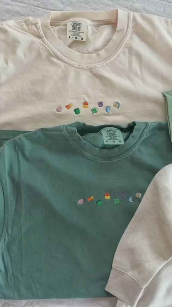Video of green and ivory adult CC tees with lucky charm marshmallows embroidered on chest and sage green child tee and natural child sweater with printed lucky charm marshmallows, seen on flat lay - Little Mama Shirt Shop