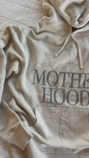 Video of adult pigment hoodie in cream with 'motherhood' printed on front with floral design behind and 'living the dream' printed on the sleeve, seen on flat lay - Little Mama Shirt Shop