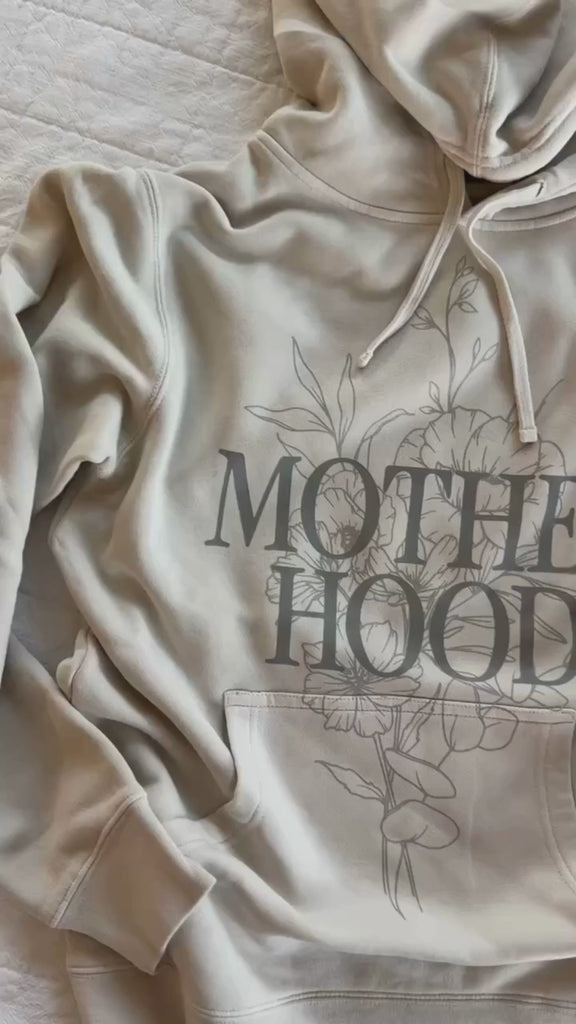 Video of adult pigment hoodie in cream with 'motherhood' printed on front with floral design behind and 'living the dream' printed on the sleeve, seen on flat lay - Little Mama Shirt Shop